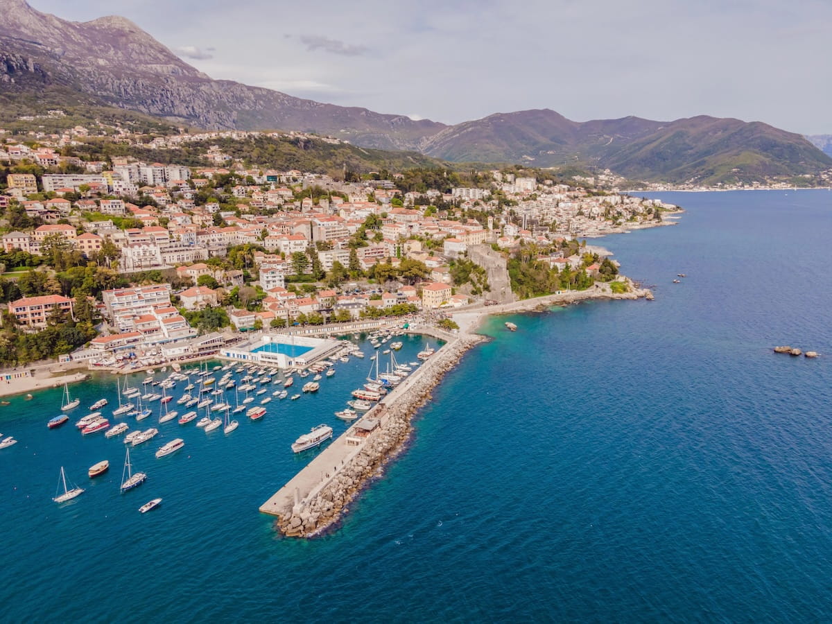 An aerial view of the coastal town of Herceg Novi, Montenegro, featuring a stone harbor pier filled with boats, a waterfront swimming pool, and red-roofed buildings climbing up a lush green hillside toward the mountains.