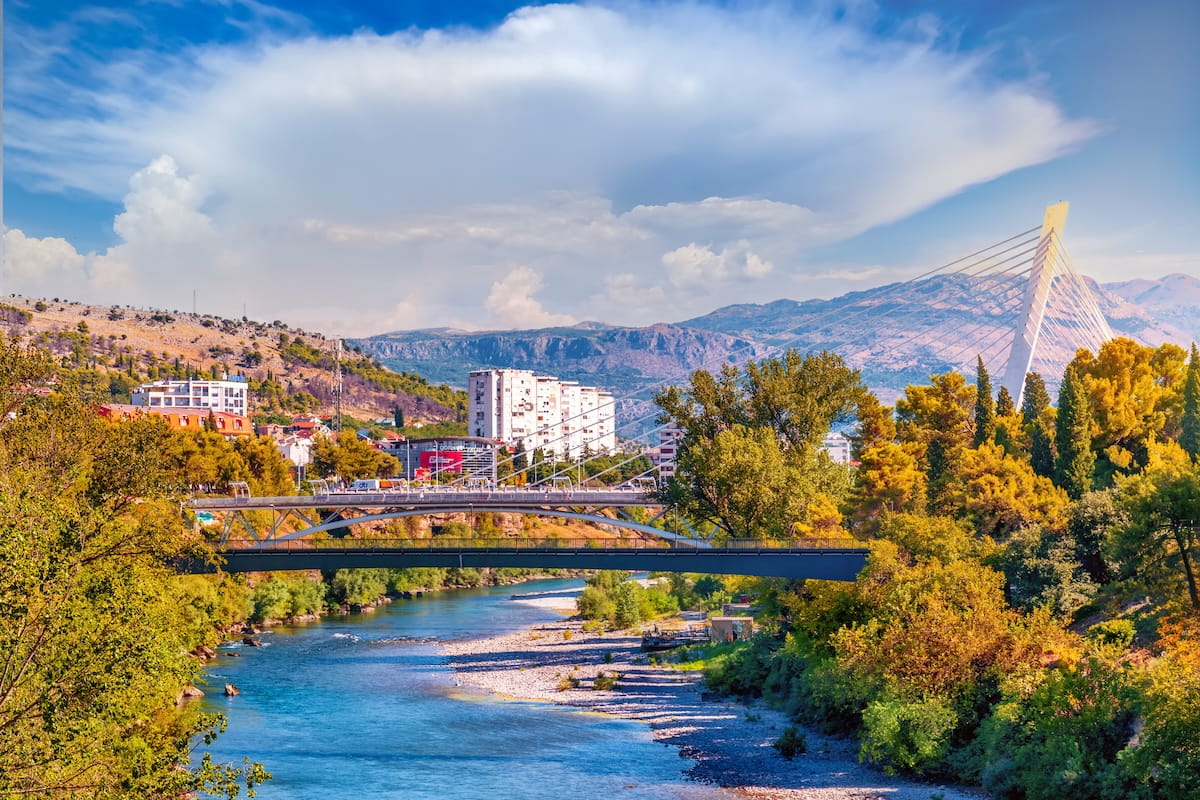 A landscape view of the white cable-stayed Millennium Bridge and the smaller Moscow Bridge crossing the blue Moraฤa River in Podgorica, surrounded by lush green and autumn-colored trees with mountains in the distance.