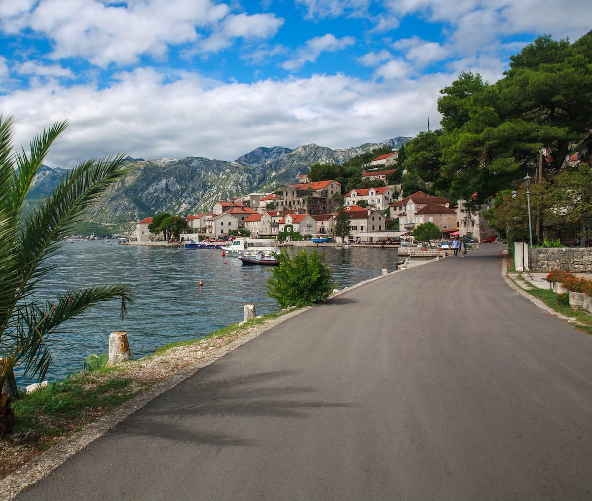 A wide-angle, scenic shot taken from a paved waterfront promenade looking towards a traditional Montenegrin coastal town in the Bay of Kotor, with the water and massive grey limestone mountains under a partly cloudy sky. The town features numerous white stone buildings with distinctive red-orange terracotta roofs clustered on the waterfront. Boats are moored in the blue-grey water, and a palm tree is visible in the foreground to the left.