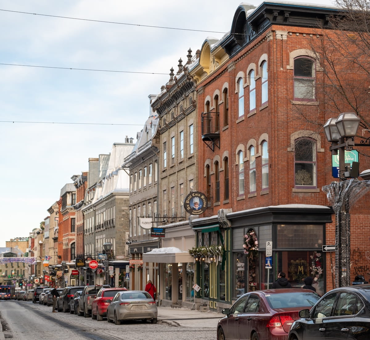 A daytime view down a historic cobblestone street in Old Québec during winter. The street is lined with multi-story European-style brick and stone buildings housing boutiques and restaurants like "Au Petit Coin Breton." Cars are parked along the side of the road, and bare trees and vintage-style lampposts are visible against a cloudy sky.