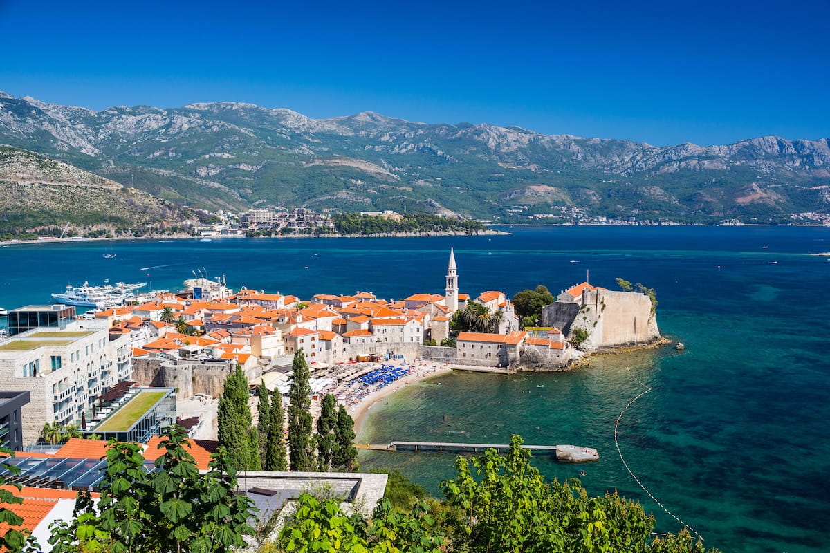 An elevated view of Budvaโs Old Town, featuring orange-roofed stone buildings, a prominent church bell tower, and the medieval Citadel fortress jutting into the turquoise Adriatic Sea against a backdrop of rugged green mountains.