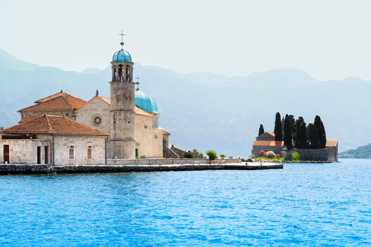 An aerial panoramic view of Our Lady of the Rocks, a man-made island with a blue-domed church sitting in the middle of the turquoise Bay of Kotor, surrounded by dark mountains.
