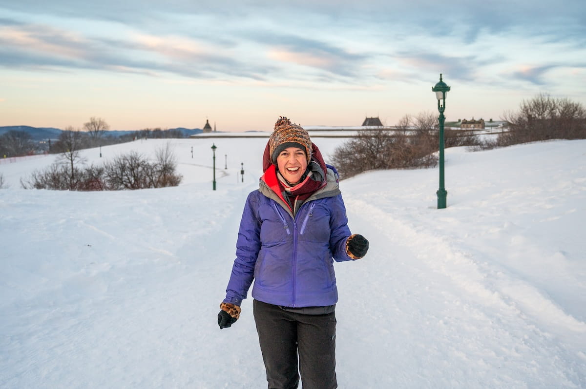 Traveler Jessie Festa smiles while walking on a wide, snow-covered path in the Plains of Abraham during winter. She is wearing a purple jacket, a knit beanie, and a scarf, with a backdrop of rolling snowy hills, historic green lampposts, and a soft sunset sky.