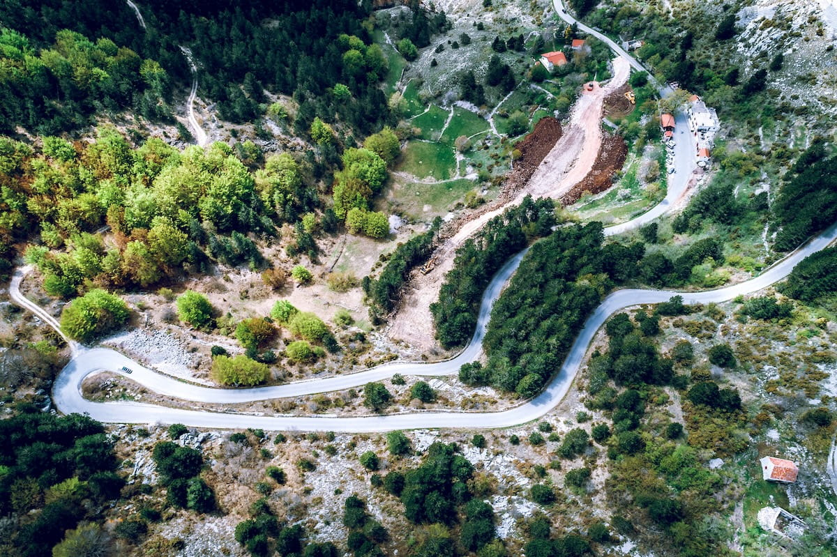 Aerial view of a winding serpentine road snaking through a lush green mountain forest in Montenegro, illustrating the challenging driving conditions for solo travelers.