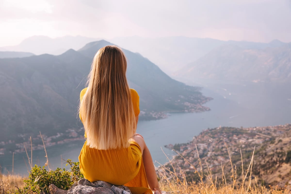 A back view of a solo female traveler with long blonde hair in a yellow dress sitting on a rocky cliff, looking out over the expansive Bay of Kotor and the hazy mountain ranges of Montenegro.