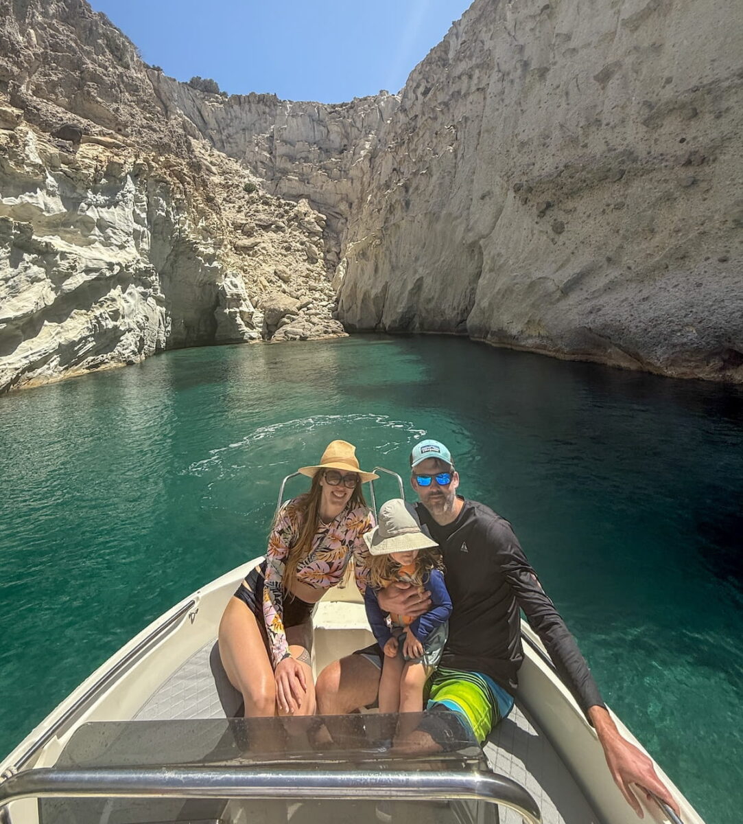 Kristin Addis and her family sitting on a small motorboat in a narrow turquoise sea canyon. Kristin is on the left in a floral rash guard and straw hat, while her husband sits on the right holding their young son. High limestone cliffs surround the clear water under a bright sun.