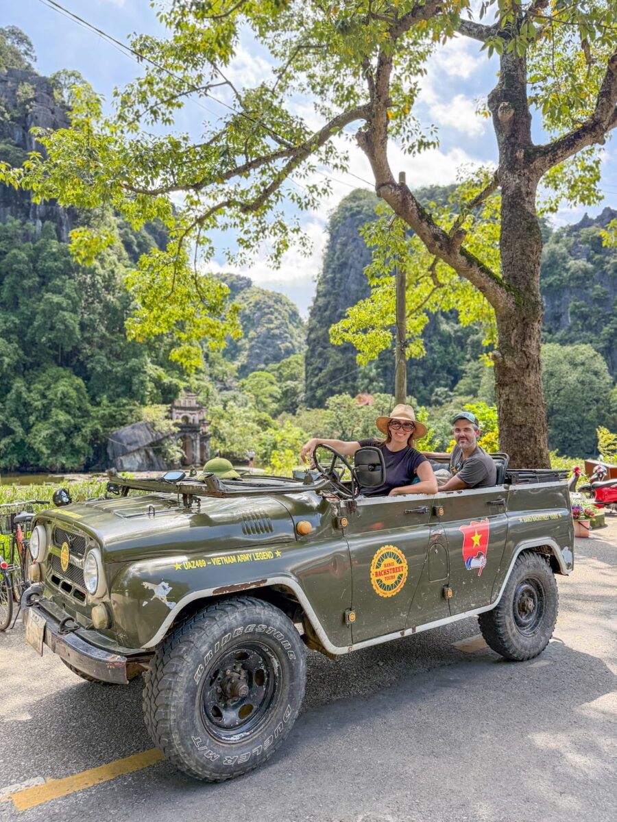 Kristin Addis and her husband sitting inside a classic olive-green military jeep (a UAZ-469 marked "Vietnam Army Legend") on an adventurous road trip. Kristin is in the driver's seat with a big smile, wearing a black top and a wide-brimmed straw hat. In the background, lush, towering limestone karsts rise above a roadway lined with vibrant green trees under a bright, partly cloudy sky.