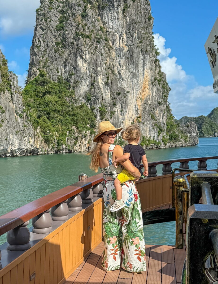 Travel creator Kristin Addis stands on the wooden deck of a boat in Ha Long Bay, Vietnam, holding her young son. She is wearing a wide-brimmed straw hat and a tropical floral print jumpsuit, looking over her shoulder with a smile. In the background, towering limestone karsts rise out of the emerald green water under a blue sky.