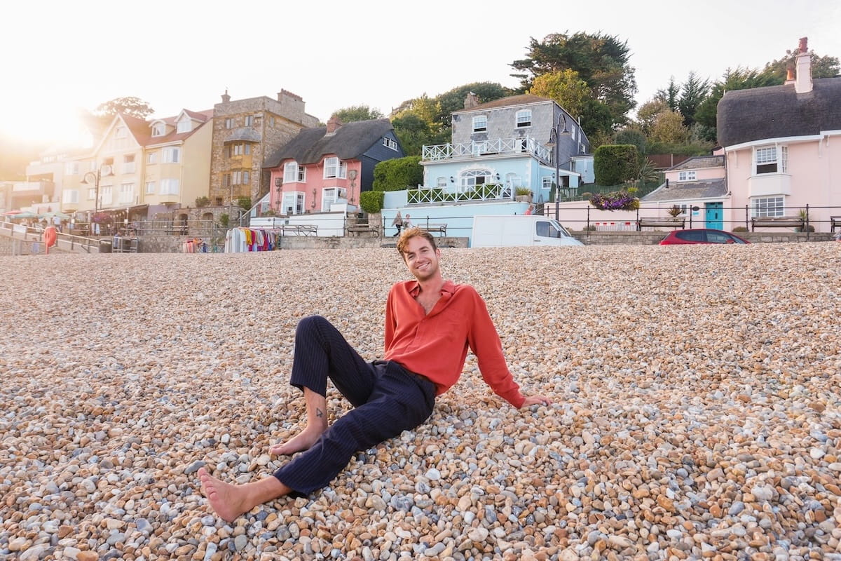 Travel influencer Kevin Droniak sits barefoot on a pebble beach, leaning back and smiling at the camera. He is wearing a burnt-orange button-down shirt and dark pinstriped trousers. In the background, a row of charming, colorful coastal houses in shades of pink, blue, and yellow sits at the base of a green hill, with the soft glow of a setting sun creating a bright flare on the left side of the frame.