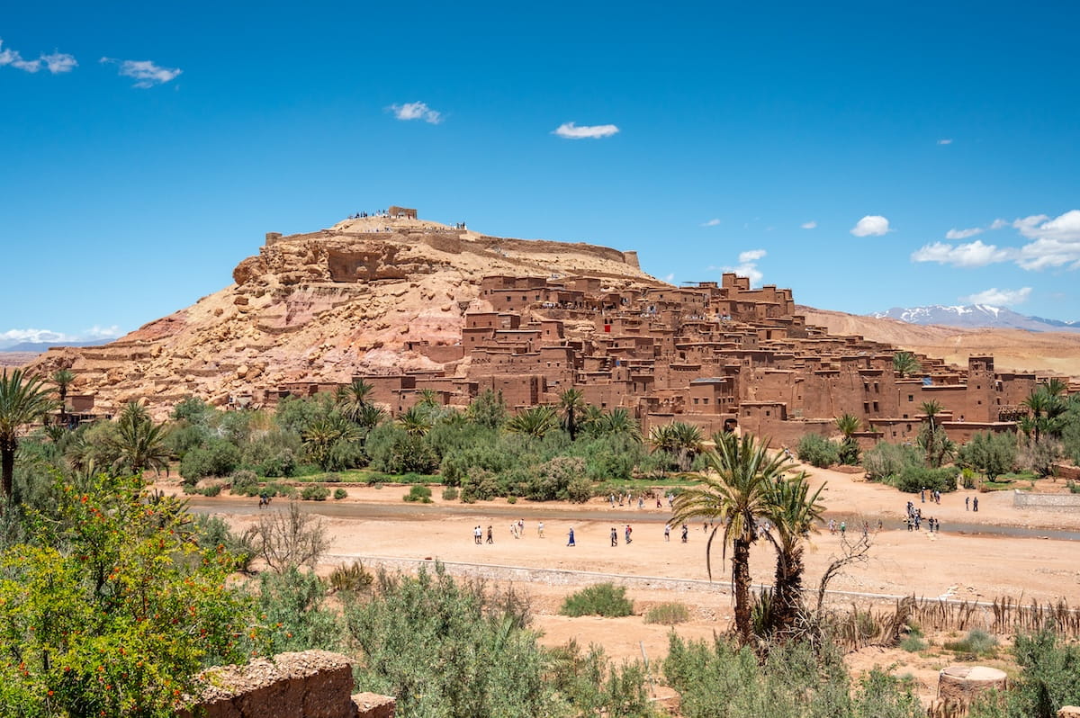 A wide, sunny landscape shot of the ancient fortified earthen village, or ksar, of Aรฏt Benhaddou in Morocco. The historic complex features tiered, reddish-brown mud-brick buildings and defensive towers built directly into the side of a large, barren hill under a vibrant blue sky. In the foreground, green palm trees and desert shrubs line a wide, dry riverbed where small groups of tourists walk along the sand, with the faint, snow-capped peaks of the High Atlas Mountains visible on the far right horizon.