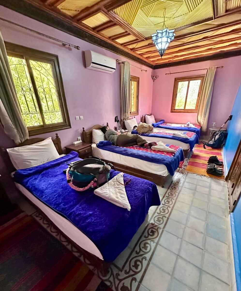 A long guest room inside the Atlas Imoula Guesthouse in Imlil, Morocco. The room features light purple walls, a traditional tiled floor border, and an intricate dark wood ceiling with a hanging metal lantern. Four single beds topped with bright blue blankets line the left wall, and a traveler lies resting on one of the beds in the center.