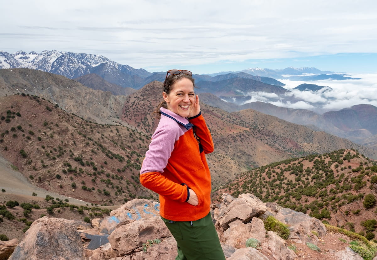 A smiling woman wearing a bright orange and pink fleece pullover stands on a rocky mountain summit. In the background, expansive views of the High Atlas Mountains unfold with layered hills, snow-capped peaks in the distance, and white clouds gathering in the valleys under a cloudy sky.