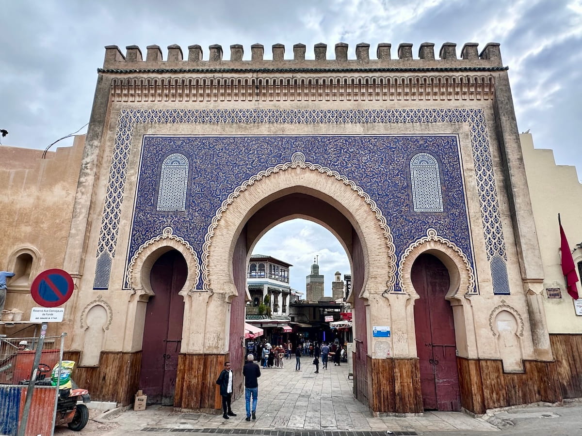 A ground-level shot of the iconic Bab Bou Jeloud (The Blue Gate), a monumental triptych archway leading into the old medina of Fes, Morocco. The massive central arch and its two flanking smaller arches are built from sand-colored stone, adorned with intricate geometric arabesque carvings and a large expanse of vibrant blue mosaic tiles surrounding the openings. The top of the gate is lined with traditional notched crenellations against an overcast sky. Pedestrians walk through the open arches into a bustling street lined with historic multi-story buildings, and a green minaret tower is visible in the distance framed perfectly by the main archway. On the far left foreground, a modern red-and-blue "no parking" traffic sign and construction fencing sit next to the stone wall.