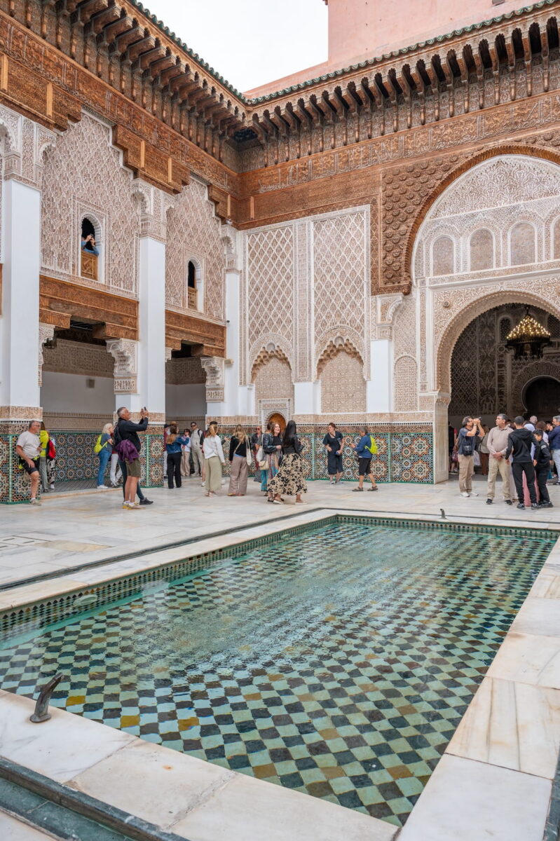 The inner courtyard of Ben Youssef Medersa in Marrakech, featuring a large, rectangular shallow reflection pool lined with green and white checkerboard tiles. Tourists wander around the white marble courtyard floor, looking at the towering walls adorned with intricate stucco carvings, geometric wood detailing, and colorful mosaic tilework. Arched doorways, carved pillars, and small upper-floor dormitory windows overlook the central pool under a bright sky.
