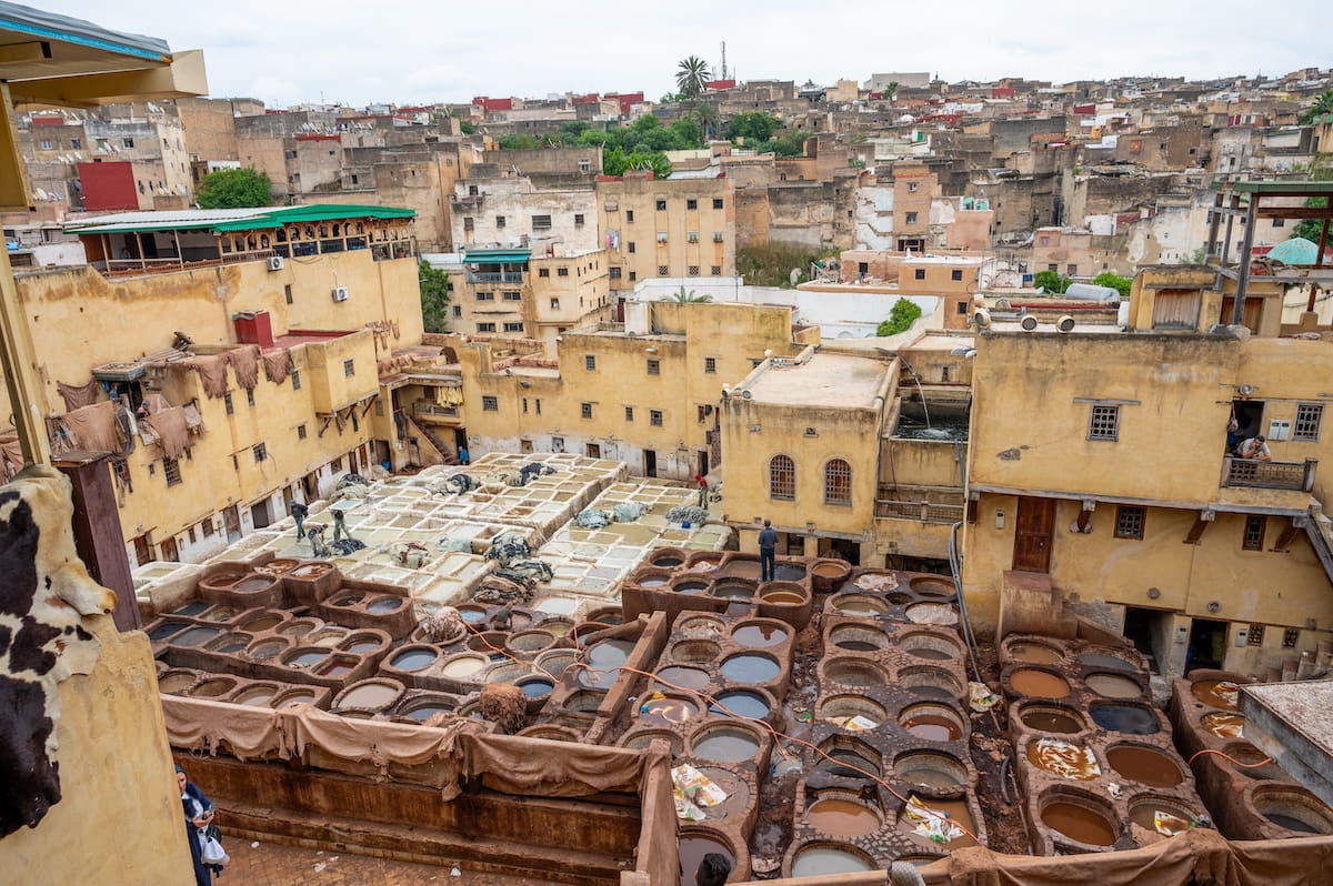 A high-angle view looking down into the expansive outdoor courtyard of the Chouara Tannery in Fes, Morocco. The historic site is filled with dozens of stone vessel pits containing various colored liquids and dyes, with workers seen tending to the hides. Surrounding the pits are multi-story, sand-colored earthen buildings, with freshly tanned animal skins draped over walls and railings to dry. In the far background, the densely packed, rolling cityscape of the Fes medina stretches toward a hazy, overcast horizon.