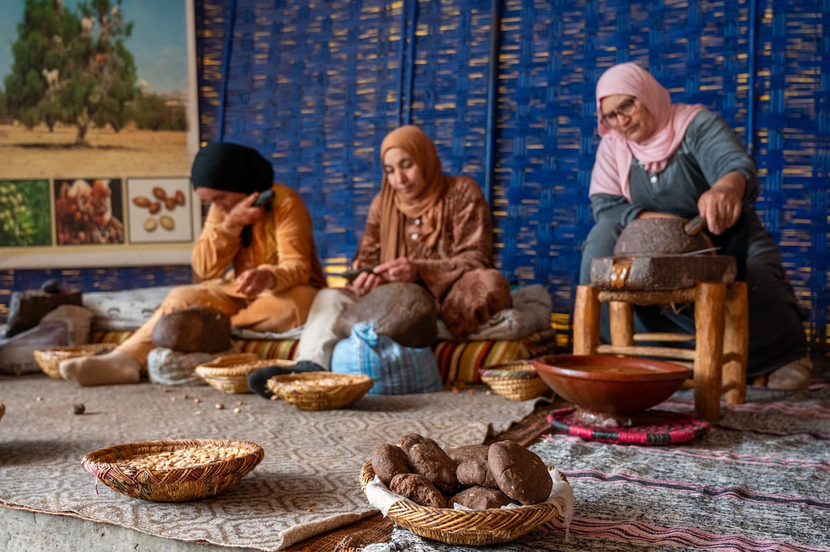 Three Moroccan women wearing headscarves sit on a carpeted floor working at an argan oil cooperative. The woman on the right uses a traditional stone grinder on a small wooden stand, while the other two women crack argan nuts in the background against a blue woven wall. Woven baskets containing nuts and argan paste paste cakes sit in the foreground.