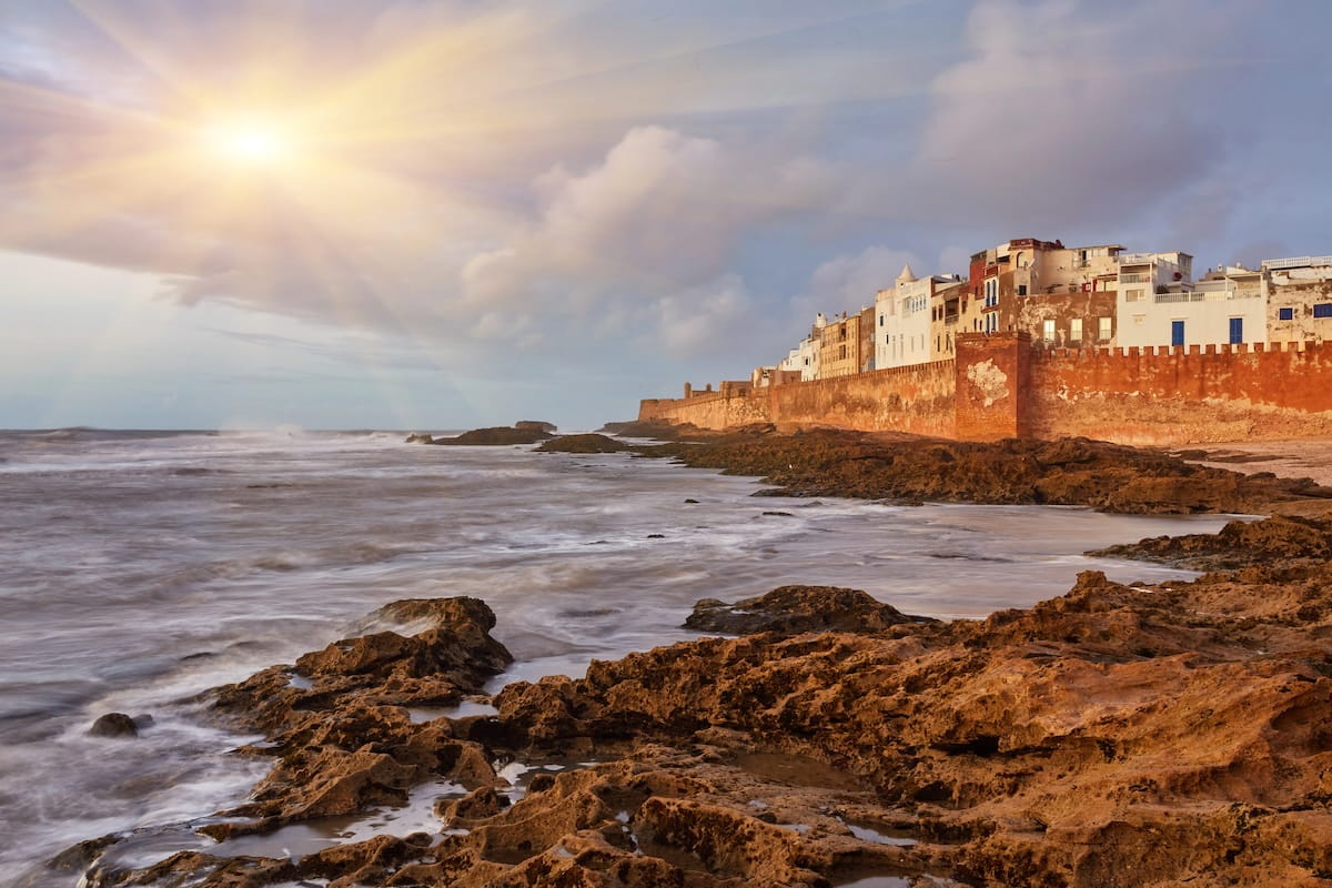 A wide landscape view of the historic fortified seaside wall and ramparts of Essaouira, Morocco, bordering the Atlantic Ocean. The foreground features a rugged, dark brown volcanic rock shoreline with water washing over the tide pools. On the right, a massive, crenellated red stone rampart stretches into the distance, supporting a row of whitewashed multi-story buildings and traditional houses. The ocean waves crash against the rocky coast on the left, while a brilliant sun bursts through dramatic, heavy grey clouds, casting golden light rays across the hazy sky.