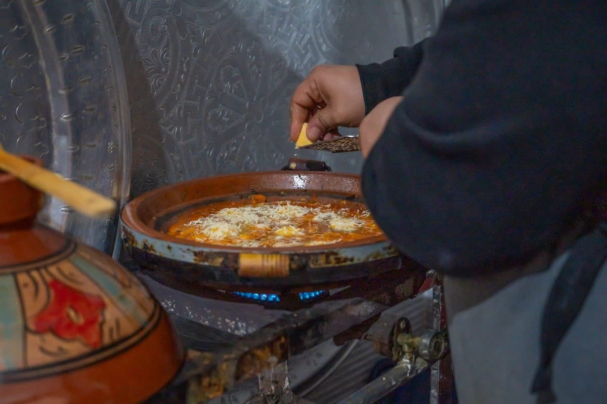 A close-up shot of a chef's hands grating cheese over a simmering dish inside a clay tagine base during a cooking class in Fes, Morocco. The tagine sits on a portable metal gas burner with visible blue flames beneath it. The dish inside features a rich, orange-red tomato sauce base topped with eggs and a layer of shredded cheese. In the blurred background, large, intricately engraved silver decorative platters stand upright, while the colorful, painted ceramic conical lid of another tagine sits in the lower left foreground.
