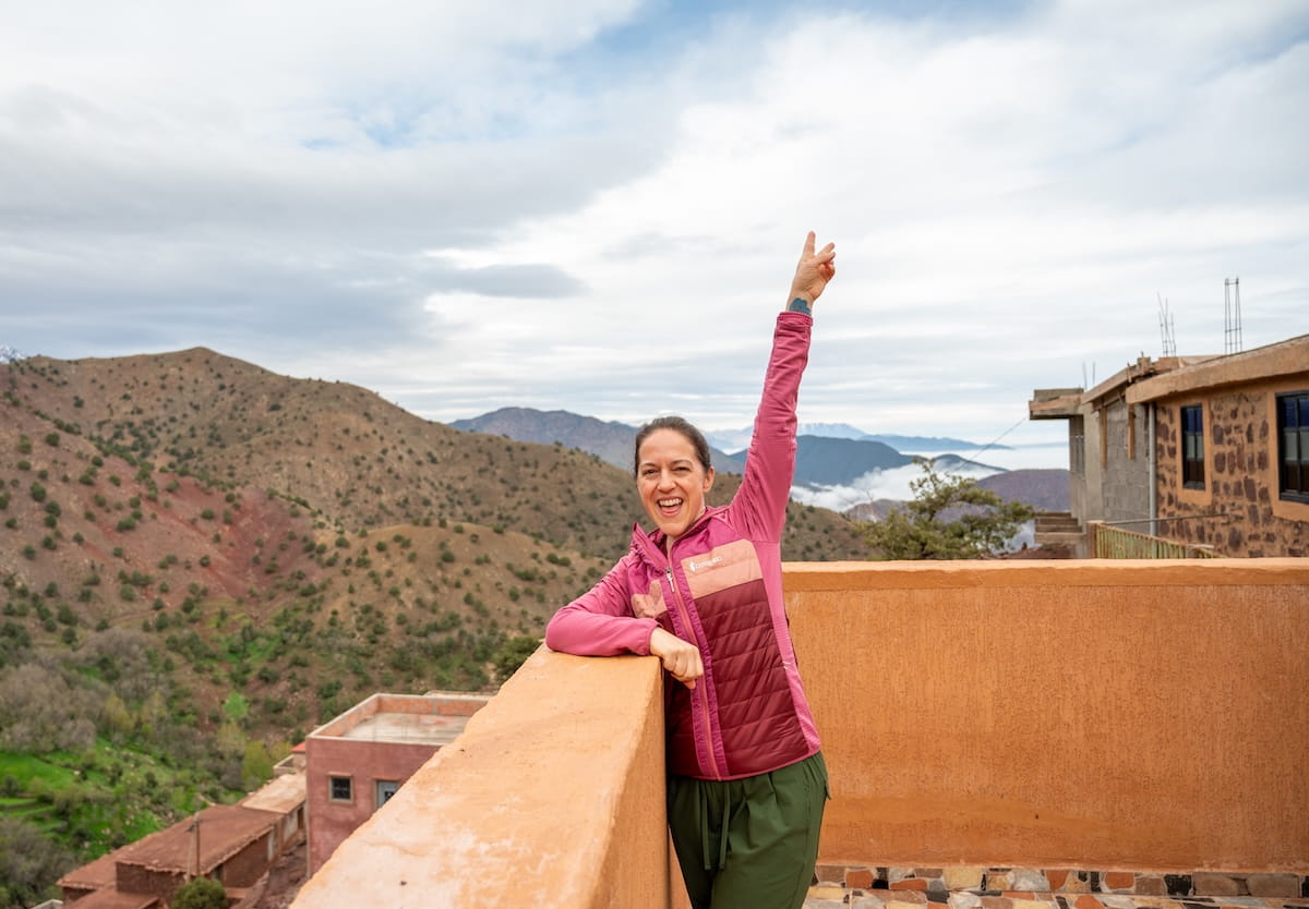 A smiling woman wearing a pink hiking jacket and green pants stands on a stone balcony, raising her arm in a peace sign. In the background, rugged mountains rise up under an overcast sky, with a thick layer of white clouds settling in the valley below.