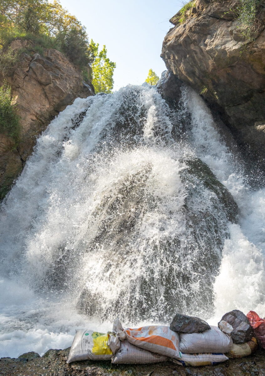 A powerful, rushing waterfall cascades down over a large dark rock formation at the Imlil Cascade in Morocco. White water sprays dramatically in all directions, framed by rocky cliffs with green trees peeking out at the top. A row of filled sandbags sits along the rocky edge in the immediate foreground.