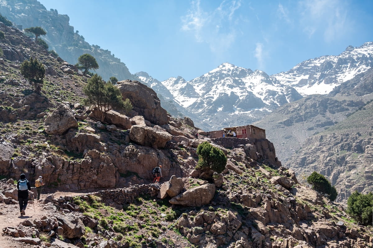 Hikers backpack up a rugged, rocky uphill trail towards a small stone mountain outpost building built into the cliffside. The steep slope is dotted with sparse green bushes and small trees, with the massive, snow-capped peaks of the High Atlas Mountains towering in the background under a bright sky.
