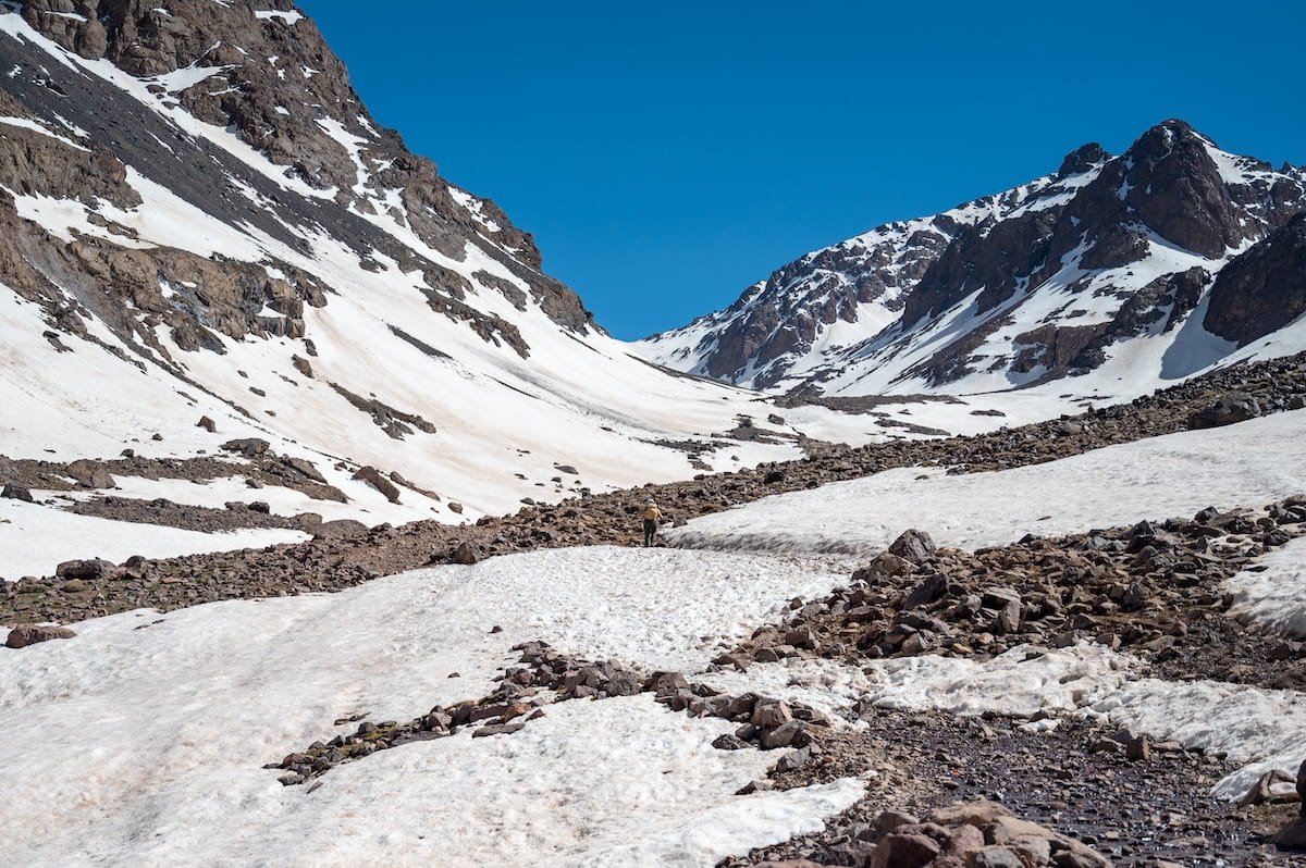 A lone hiker walks up a vast, snow-covered valley on the trek from Imlil to the Toubkal Refuge in Morocco's High Atlas Mountains. Large, rocky brown mountains patches with snow rise sharply on both sides of the snowy trail under a clear, bright blue sky.