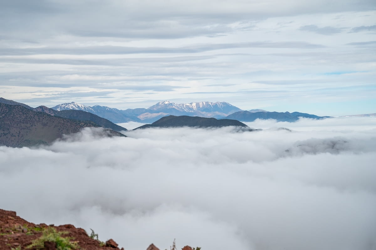 A breathtaking panoramic view of the High Atlas Mountains with a thick blanket of white clouds completely filling the valley floor. Layered, dark mountain ridges cut through the sea of clouds, while snow-capped peaks rise in the far distance under a dramatic, overcast gray sky.