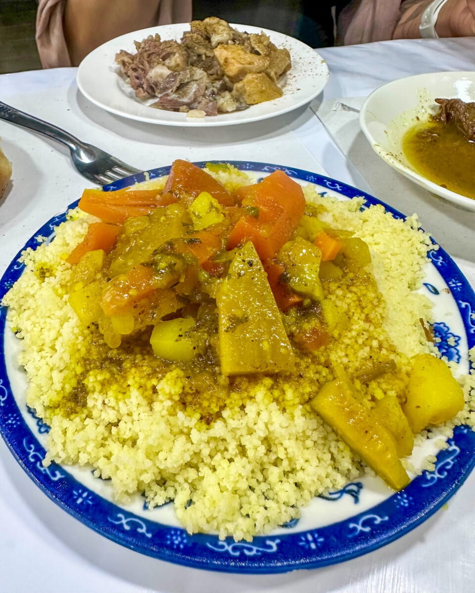 A large plate of vegetable couscous topped with cooked carrots, potatoes, and squash in a savory sauce from Chez Hassan in Marrakech. The plate is white with a blue decorative rim and sits on a table alongside other dishes of food.