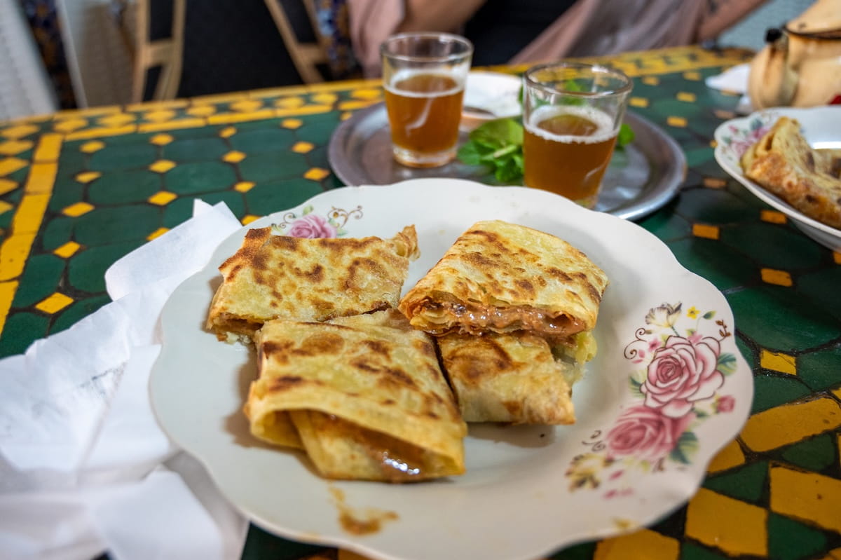 A plate of folded Moroccan msemen pancakes cut into pieces on a decorative floral plate. In the background, two small glasses of mint tea sit on a metal tray on top of a green and yellow tiled table.