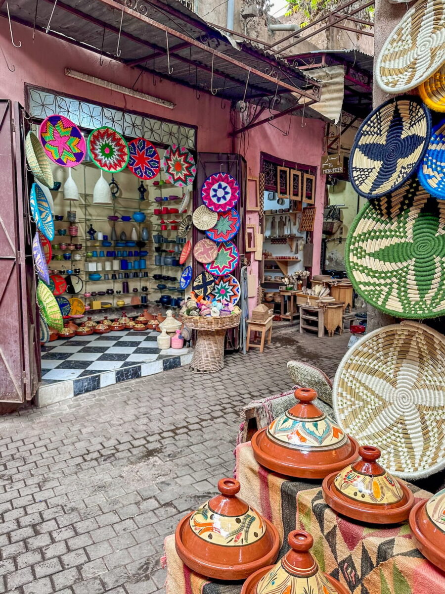 An eye-level shot capturing a vibrant alleyway shop in the Marrakech Medina, Morocco, filled with traditional handicrafts. The cobblestone pathway leads past an open-front storefront with pink stucco walls and a black-and-white checkered tile floor inside, its shelves tightly packed with colorful ceramics, cups, and pottery. Hanging prominently around the doorway and a neighboring wooden pillar are numerous hand-woven decorative baskets and platters showcasing bold, geometric star patterns in neon pink, green, blue, yellow, and red. In the right foreground, several glazed ceramic tagines adorned with painted details sit on a draped woven blanket, while a glimpse of a woodworking shop with wooden utensils is visible down the narrow corridor in the background.