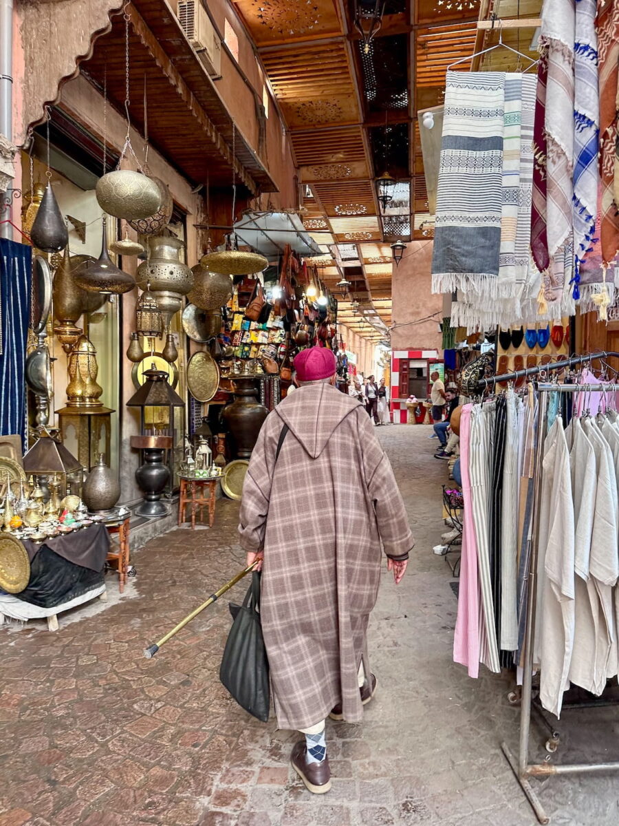 A man wearing a traditional plaid djellaba robe and a red fez hat walks down a narrow covered alley in the Marrakech Medina. Hanging brass lamps and lanterns line the shops on the left, while clothing racks with textiles and robes line the right.