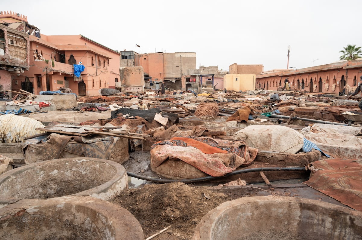 A ground-level view looking across the expansive outdoor courtyard of the Marrakech Tanneries in Morocco. The foreground is dominated by large, circular stone pits stained with mud and dyes, some containing dark liquid and others draped with thick, heavy cloths. Throughout the wide courtyard, piles of raw hides, bundled leather skins, and patterned textiles sit drying on flat surfaces and stone structures under a hazy, overcast sky. Workers can be seen in the mid-ground managing the materials. The space is enclosed by low, pinkish-orange stucco buildings featuring traditional arched windows, with a single palm tree visible on the far right horizon.