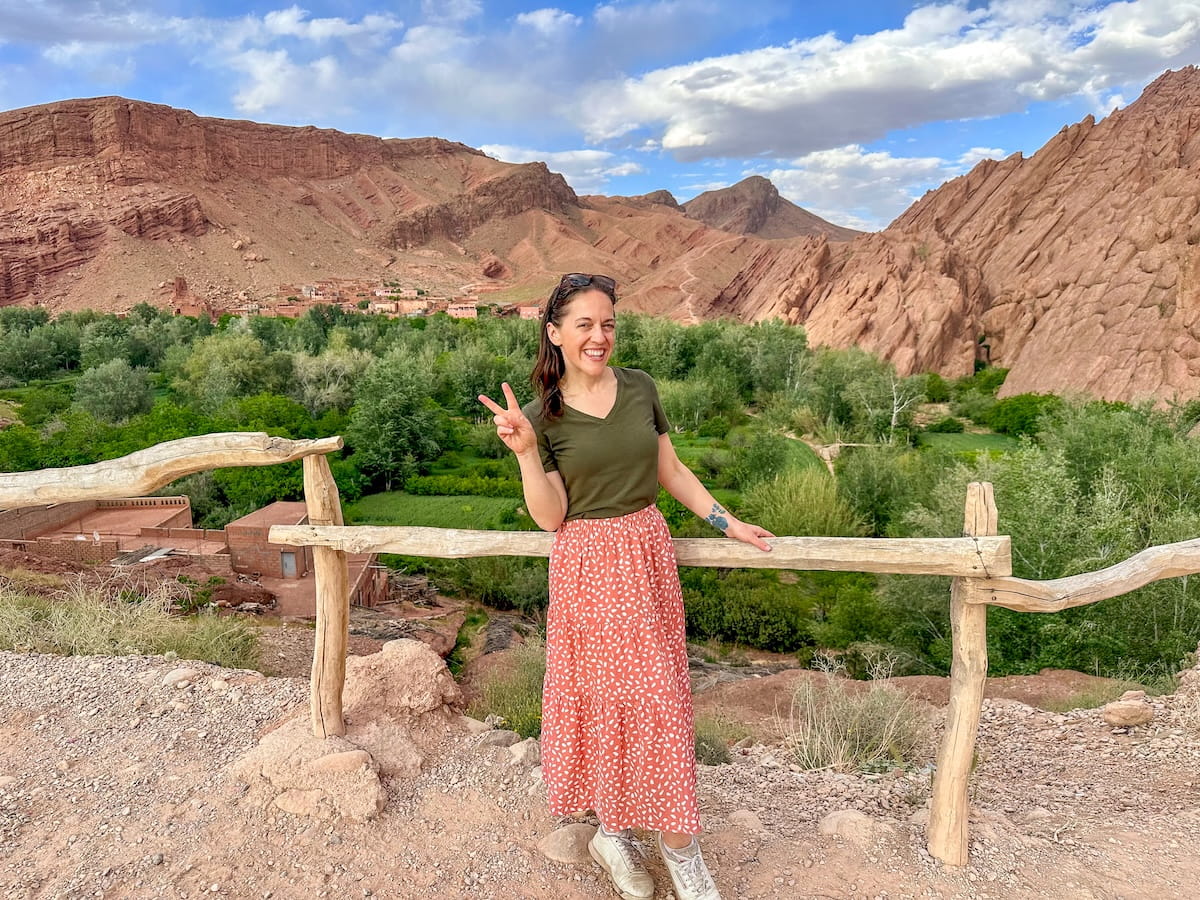 A smiling woman wearing an olive green v-neck t-shirt and a long, patterned pink and white maxi skirt stands behind a rustic wooden log railing at the Monkey Fingers Lookout in Morocco's Dades Gorge. She has sunglasses perched on her head and flashes a peace sign with her right hand. Behind her, a vibrant green valley filled with dense trees and agricultural fields stretches across the mid-ground, leading to a small village nestled at the base of massive, uniquely eroded red rock formations under a blue sky with soft white clouds.