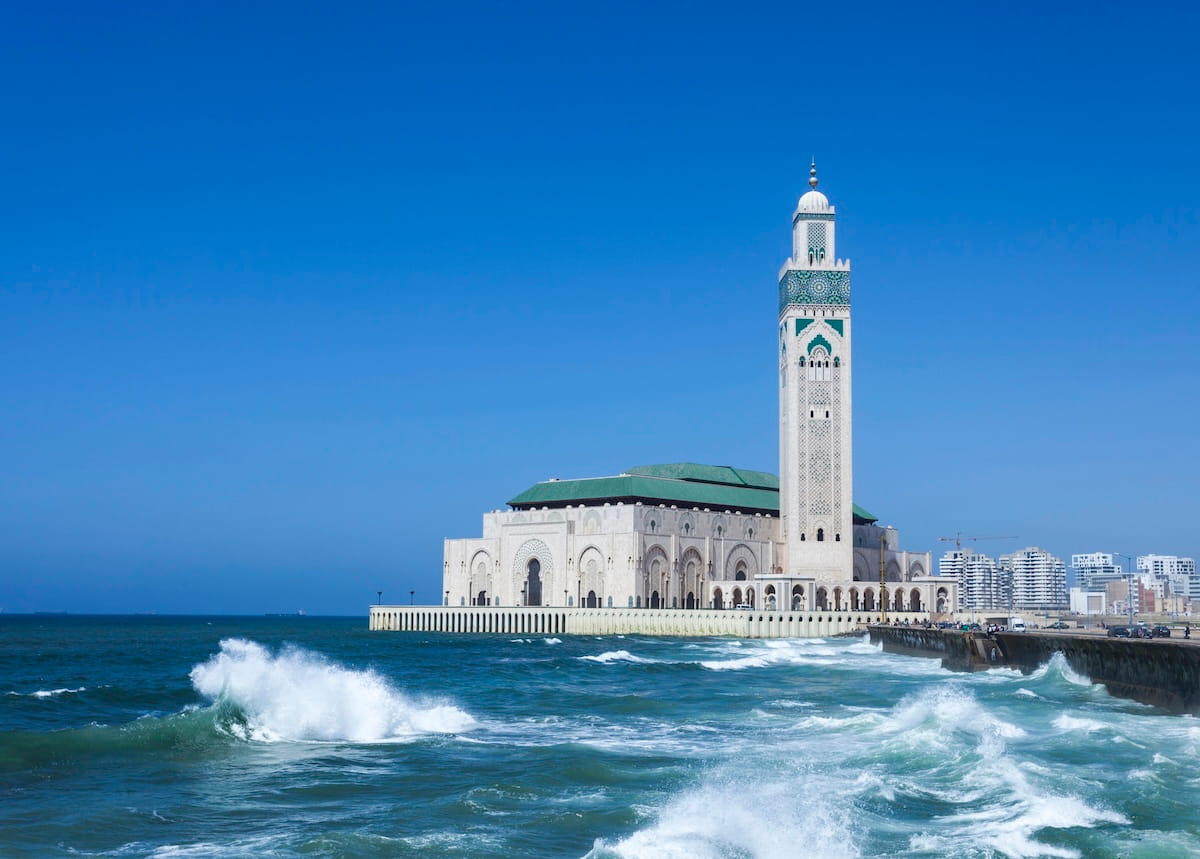 A wide-angle landscape shot of the grand Hassan II Mosque in Casablanca, Morocco, built on a platform directly over the Atlantic Ocean. The massive cream-colored stone complex features intricate arched doorways, a green-tiled roof, and a soaring minaret tower adorned with green geometric mosaic work pointing into a clear, deep blue sky. In the foreground, powerful cerulean ocean waves topped with dramatic white seafoam crash aggressively against a stone seawall on the right, where a few distant figures can be seen walking.