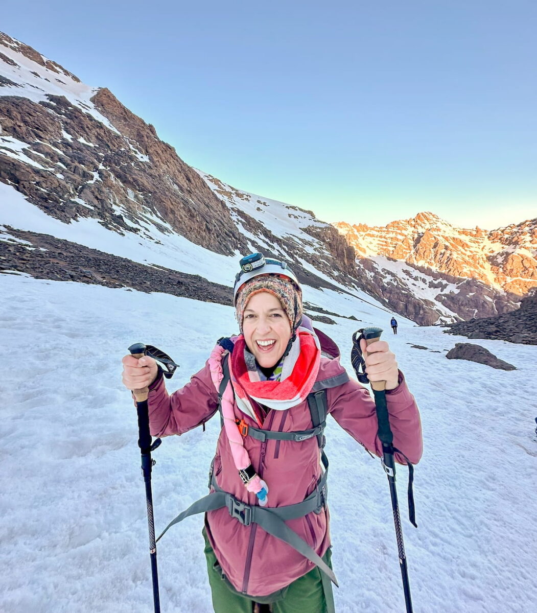 A close-up, front-facing shot of a smiling woman hiking on a snow-covered slope during an early morning trek up Mount Toubkal in Morocco's High Atlas Mountains. She wears a helmet with a headlamp, a multi-colored knit beanie, a bright red and white scarf, a purple jacket, and green pants while holding up two black trekking poles. The steep snowy trail behind her shows a distant hiker, flanked by dark rocky mountainsides on the left and sunlit, orange-tinted peaks in the background under a pale morning sky.