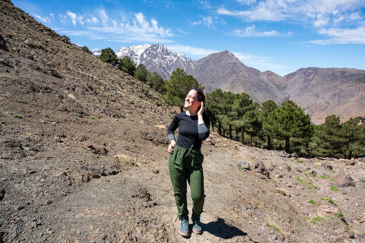 A smiling woman wearing an orange and pink fleece pullover stands on a rocky trail at the Tizi n' Tmatrt pass in Morocco's High Atlas Mountains. In the background, expansive views of rugged valley ridges and distant snow-capped peaks unfold under a cloudy sky.