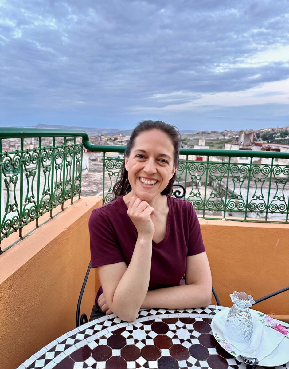 A close-up shot of a smiling woman sitting at a mosaic tile table on the rooftop terrace of Restaurant Dar Trรฉsor in Fes, Morocco. She is wearing a maroon v-neck t-shirt and rests her chin thoughtfully on her right hand. The circular table in front of her features an intricate brown and white star-patterned tile design, set with an upturned clear glass resting on a white napkin and plate. Behind her, a low orange stucco wall is topped with an ornate green iron railing, offering a sweeping view of the sprawling city of Fes stretching out beneath a vast, cloudy evening sky.