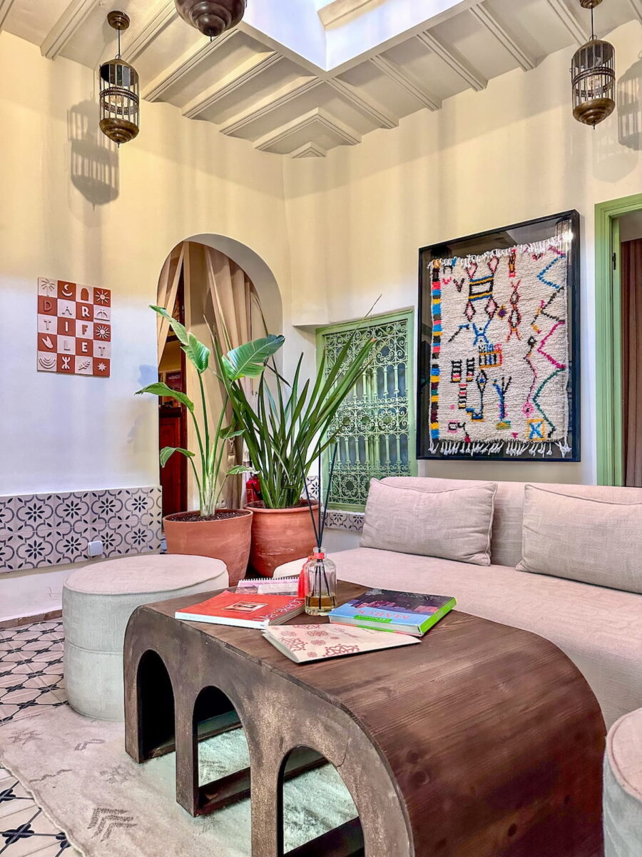 Sunlit lounge courtyard at Riad Dar Tiflet in Marrakech featuring a sofa, woven rug wall hanging, and potted plants.
