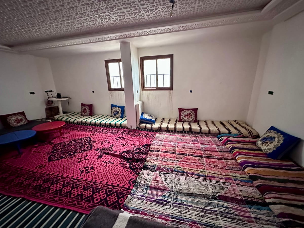 A large, traditional Moroccan room lined with low cushion seating, patterned bolsters, and colorful woven rugs on the floor. Two small windows look out from the white walls beneath an intricately textured white ceiling.