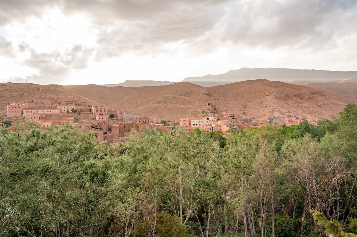 A scenic rooftop view looking out across a dense grove of green trees toward a traditional Moroccan hillside village in the Dades Gorge. The earthen, reddish-brown buildings are nestled along the slopes of barren, rolling hills under a soft, overcast sky with sunlight breaking through the clouds.