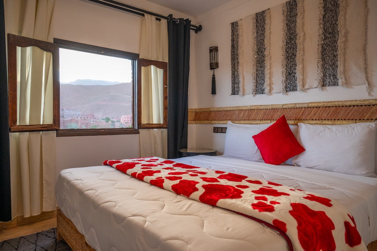 A bright bedroom inside a riad at the Dades Gorge, featuring a large bed made up with crisp white linens, a bright red accent pillow, and a matching red-and-white patterned blanket. An open window with dark curtains and wooden shutters looks out onto the arid mountain landscape, while a woven black-and-white textile wall hanging completes the room's traditional decor.