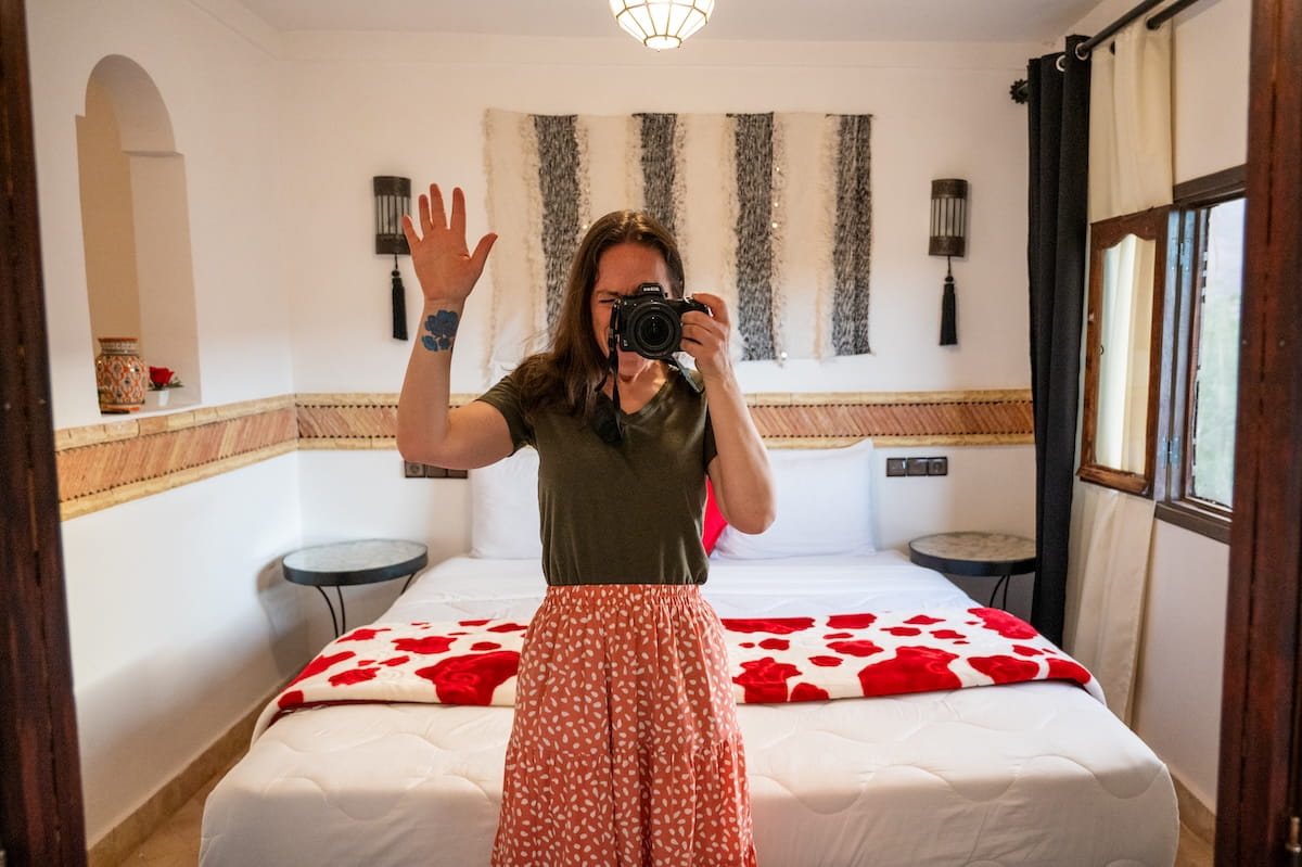 A mirror-reflection selfie of a woman showing a conservative travel outfit in her room at a riad in the Dades Valley, Morocco. She stands in the center of the frame holding a black DSLR camera up to her eye to take the photo while raising her right handโrevealing a blue floral tattoo on her inner wristโin a small wave. She is wearing an olive green v-neck t-shirt tucked into a high-waisted, patterned pink-and-white maxi skirt. The background reflects the hotel room, featuring a large bed with crisp white linens, a bright red-and-white patterned blanket, and a traditional black-and-white woven textile hanging on the wall above the headboard.