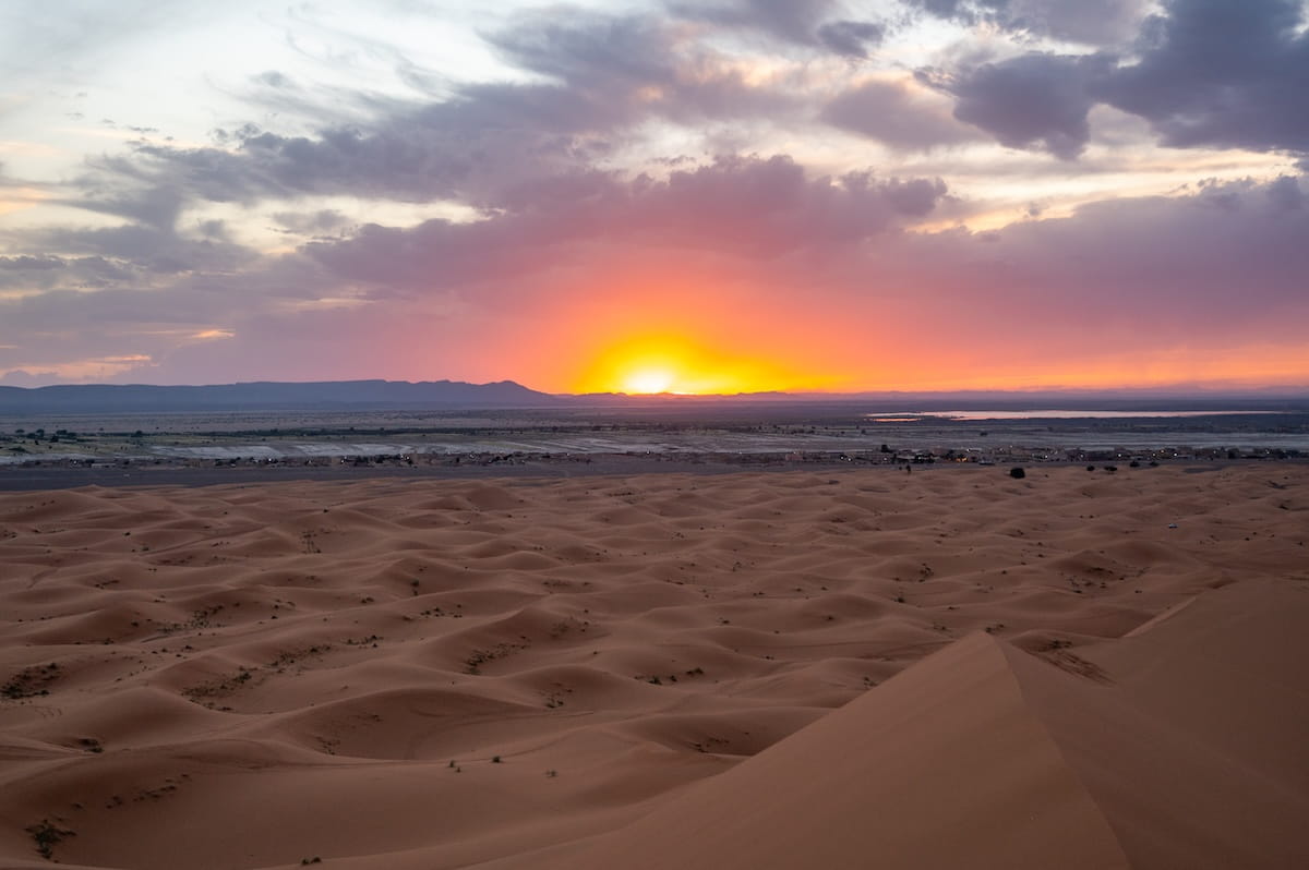 A high-angle panoramic view of a vibrant sunset over the Sahara Desert in Morocco. In the foreground, vast undulating patterns of orange sand dunes stretch across the landscape, with a sharp dune ridge sloping down on the right. In the mid-ground, a low-lying desert village sits flat against a barren plain, with a glimpse of a lake visible on the far right. The sun sinks behind dark, flat-topped mountain ridges on the horizon, illuminating the dramatic cloud cover above in brilliant shades of bright orange, pink, and deep purple.