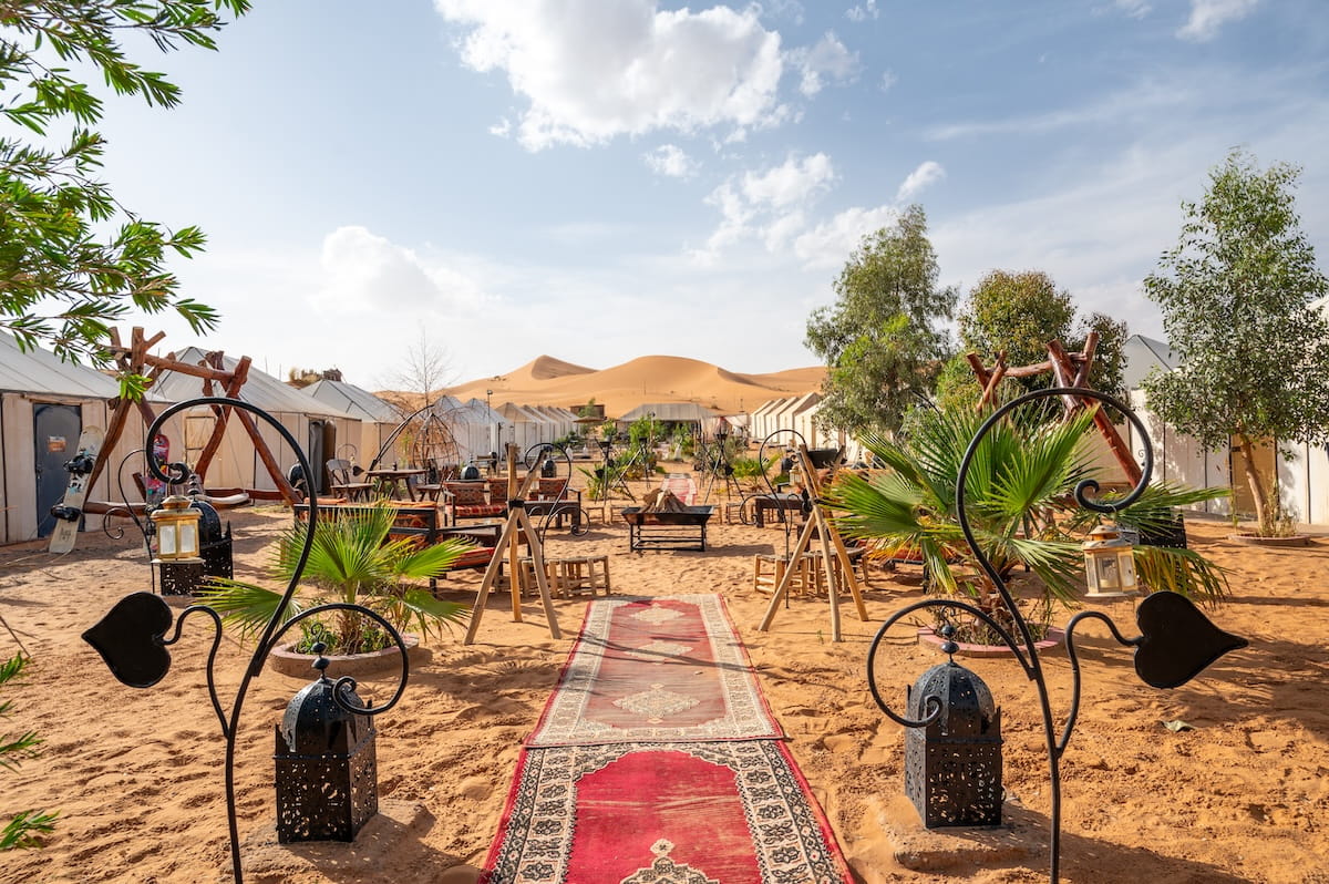 A wide shot of the central sandy courtyard at the Sanmao Desert Luxury Camp in the Sahara Desert, Morocco. A long red patterned rug runner forms a walkway through the sand in the foreground, flanked by two ornate black metal lanterns on stone bases. The open-air courtyard features seating areas with patterned cushions, wooden swings, small palm trees, and a central fire pit. Lines of white luxury canvas tents stretch down both sides of the camp, leading toward a massive, smooth orange sand dune rising under a bright blue sky with light clouds.