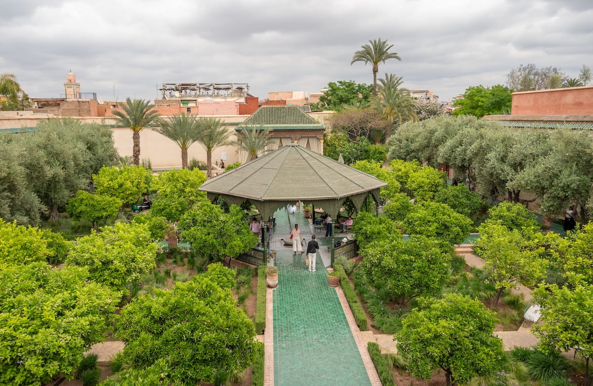 An elevated, wide-angle view of Le Jardin Secret courtyard in Marrakech under an overcast sky. A long path made of green zellij tiles leads directly to a large, open-air wooden gazebo with a green-shingled octagonal roof where a few tourists are standing. Lush greenery, manicured hedges, and mature olive and citrus trees fill the gardens on both sides of the path. In the background, sand-colored medina walls, palm trees, and a distant mosque minaret are visible.