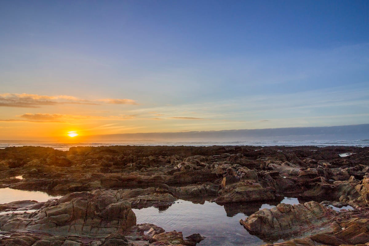 A wide-angle landscape shot capturing a golden sunset over the rugged coastline of Dar Bouazza beach in Tamarist near Casablanca, Morocco. The foreground is filled with textured, dark volcanic rock formations and small tide pools reflecting the sky. In the mid-ground, the low-hanging sun sinks toward the Atlantic Ocean horizon, casting a bright orange and yellow glow across a thin band of low clouds. The upper portion of the frame features a vast, clear blue gradient sky fading into soft evening tones.