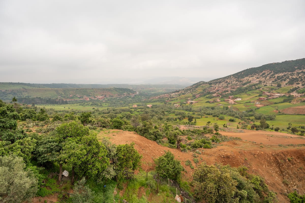 A high-angle panoramic view of a lush, rolling green valley in Morocco taken from the Tiziait Barka Cafe. The foreground features terraced red-dirt hillsides dotted with thick green trees and shrubs. In the mid-ground, a small village of traditional earthen houses is scattered across the green fields of the valley floor, leading toward rolling hills under a heavy, overcast grey sky.
