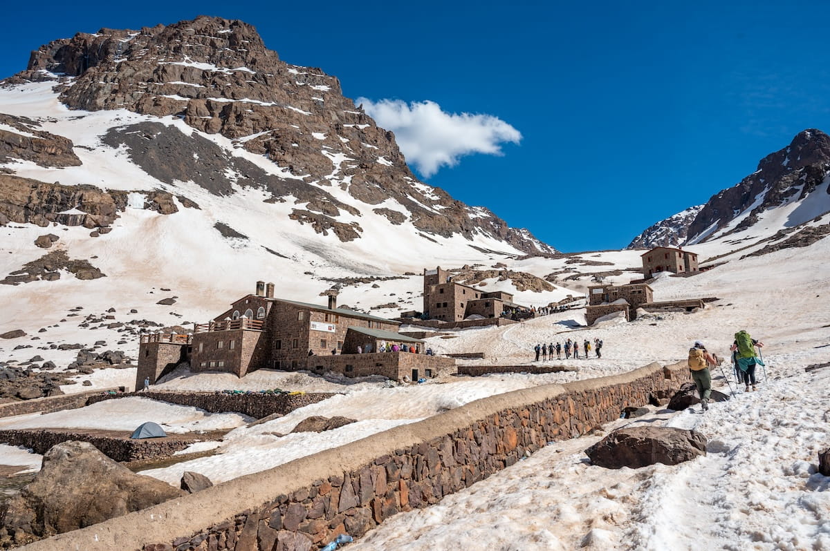 A wide landscape view of the stone buildings making up the Toubkal Refuge complex nestled at the base of a massive mountain peak in Morocco's High Atlas Mountains. The entire valley is covered in white snow, save for a long, low stone retaining wall cutting horizontally across the foreground. Hikers with backpacks and trekking poles follow a snow trail on the right toward the stone complex, under a deep blue sky with a single white cloud.