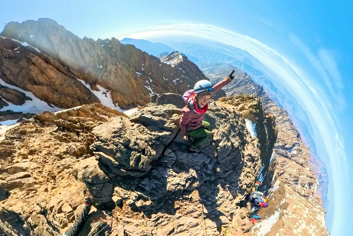 A fish-eye lens panoramic view from the rocky summit of Mount Toubkal in Morocco. A woman wearing a helmet, a red scarf, a purple jacket, and green pants sits perched on a jagged crag at the peak, smiling and pointing toward the horizon. The curved, spherical perspective captures steep rock faces dusted with patches of snow dropping off on the left, with countless layered ridges of the High Atlas Mountains stretching out toward the horizon under a bright blue sky.