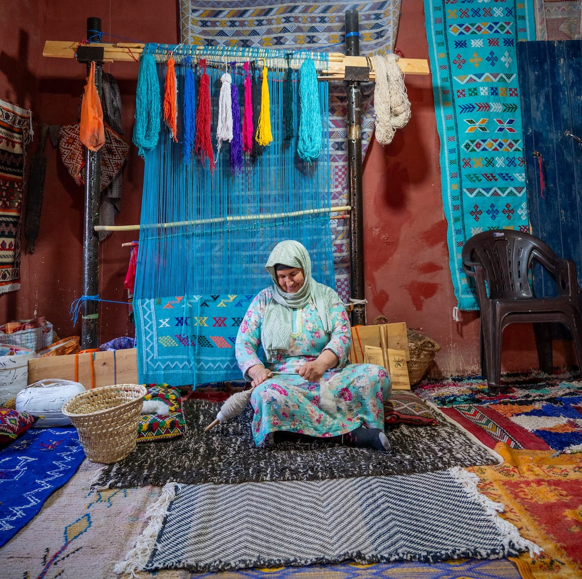 A Moroccan woman wearing a light blue and pink floral kaftan and a light green hijab sits cross-legged on a patterned rug at the Women's Carpet Association in the Todra Valley. She is smiling down as she works with a wooden drop spindle to spin raw wool into yarn. Behind her stands a large wooden weaving loom strung with bright blue vertical threads, adorned with hanging bundles of vibrant orange, red, purple, yellow, and blue yarn. The surrounding room features reddish-brown earthen walls lined with completed, colorful woven rugs, a dark plastic chair to the right, and a woven basket sitting on the floor to her left.